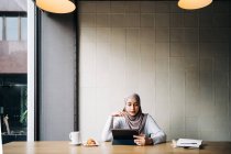 Hembra musulmana en hiyab y hablando en video chat a través de una tableta mientras está sentada en la mesa en la cafetería - foto de stock