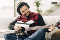 Content adult male artist in headset playing bass guitar on couch in house room in daylight — Stock Photo
