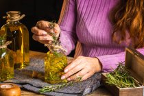 Crop señora anónima en suéter púrpura que muestra botellas de vidrio de aceite esencial con ramitas de hierbas con hojas verdes cerca de tela en la mesa - foto de stock