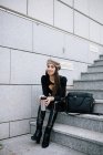 Восхитительная стильная предпринимательница в модном наряде и беретной шляпе сидит на лестнице с напитком, чтобы пойти в город и отвести взгляд — стоковое фото