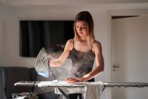 Frontansicht einer jungen Frau, die an einem sonnigen Tag zu Hause weißes Hemd bügelt, während das Bügeleisen dampft — Stockfoto