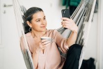 Mujer positiva de mediana edad con taza de bebida caliente que tiene video chat en el teléfono inteligente mientras está sentado en una cómoda hamaca en casa sobre un fondo borroso - foto de stock
