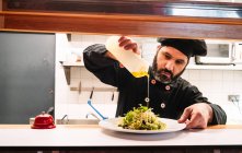 Сосредоточенный мужчина-повар в униформе выливает соус на аппетитное блюдо на тарелку во время работы на кухне ресторана — стоковое фото