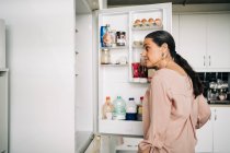 Vista lateral da fêmea calma com porta de abertura de rabo de cavalo da geladeira com vários produtos enquanto está em pé na cozinha com armários brancos — Fotografia de Stock