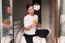 Contenuto giovane donna in abiti casual bere succo d'arancia fresco e messaggistica su smartphone mentre riposa sul letto alla luce del giorno — Foto stock