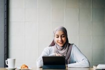 Conteúdo Mulher muçulmana no hijab e conversando no chat por vídeo via tablet enquanto se senta à mesa no café — Fotografia de Stock
