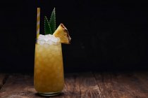 Holztisch mit einem Glas gelbem Cocktail mit Eiswürfeln und erfrischendem Cocktail garniert mit stacheligen Blättern und gestreiftem Stroh — Stockfoto
