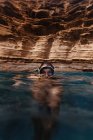 Mulher viajante em máscara nadando em água azul limpa contra penhasco rochoso durante a viagem olhando para a câmera — Fotografia de Stock