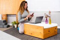 Zufriedene junge Frau mit langen Haaren in lässiger Kleidung, die Nachrichten auf dem Smartphone sendet, während sie aus der Ferne am Laptop am Boden an einem kleinen Holztisch in einer minimalistischen Wohnung sitzt — Stockfoto
