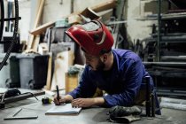 Travailleur masculin barbu sérieux en casque dur et prenant généralement des notes dans un cahier avec stylo sur la table près de constructions métalliques dans un garage léger — Photo de stock
