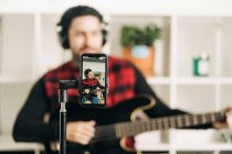 Trípode con pantalla de teléfono celular que representa la fotografía del músico masculino en auriculares tocando el bajo en casa - foto de stock