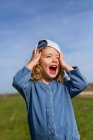 Счастливая блондинка в шапке касается головы и смотрит прочь с открытым ртом против голубого неба летом на лугу — стоковое фото