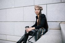 Side view of delighted stylish female entrepreneur in trendy outfit and beret hat sitting on stairs with beverage to go in city and looking away — Stock Photo