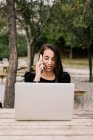 Жінка-підприємець сидить за столом з ноутбуком в парку і виступає на смартфоні під час роботи віддалено — стокове фото