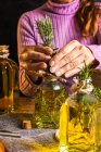 Безликая женщина в фиолетовом свитере, кладет травяные веточки с зелеными листьями в бутылку из-под эфирного масла рядом с ножницами и веревку с маленькой грудью на ткани за деревянным столом — стоковое фото