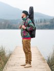 Side view of thoughtful male in stylish clothes standing with guitar in case on wooden pier near calm river against mountain in cloudy day — Stock Photo
