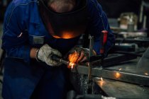 Безликий работник в перчатках и унифицированных сварных металлических деталях на столе рядом со строениями на заводе — стоковое фото