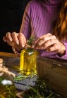 Crop anonymous lady in purple sweater showing essential oil glass bottles with herbs sprigs with green leaves near cloth on table — Stock Photo