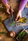 De cima da senhora de colheita cortando raminhos de alecrim com tesoura à mesa com tecido e corda perto de garrafas de vidro de óleo essencial e peito pequeno — Fotografia de Stock
