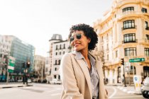 Jovem afro-americana feliz em roupa elegante sorrindo e olhando para longe na rua urbana em sol — Fotografia de Stock