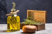 Bottiglia di vetro di olio essenziale con ramoscelli di rosmarino e candela di legno organico bruciante su tavolo grigio — Foto stock