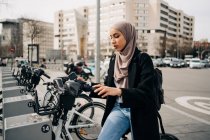 Vista lateral da mulher muçulmana em lenço de cabeça usando o sistema de compartilhamento de bicicletas na cidade — Fotografia de Stock