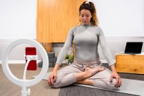 Ganzkörper einer konzentrierten jungen Frau in Aktivkleidung, die mit geschlossenen Augen in Lotus-Pose sitzt und während einer Yoga-Sitzung zu Hause in der Nähe des Smartphones meditiert, das auf einem Stativ steht — Stockfoto