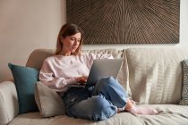 Все тело мирной молодой женщины фрилансер в повседневной одежде сидит на уютном диване и работает дистанционно на ноутбуке — стоковое фото