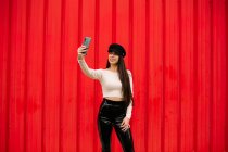Cheerful trendy female entrepreneur standing against red wall in city and taking self shot on smartphone — Stock Photo