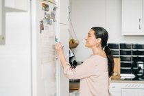 Vue latérale de la femme d'âge moyen positif prenant des notes dans le calendrier sur le réfrigérateur tout en ayant un appel téléphonique dans la cuisine moderne — Photo de stock