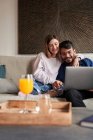 Веселая молодая многонациональная пара в повседневных нарядах, улыбающаяся сидя на диване и разговаривая на видео через ноутбук — стоковое фото