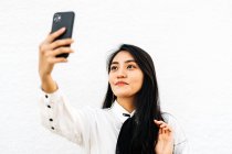 Asiatin mit langen dunklen Haaren fotografiert Selbstporträt auf Handy stehend auf weißem Hintergrund — Stockfoto