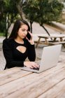 Женщина-предприниматель сидит за столом с ноутбуком в парке и говорит на смартфоне во время работы удаленно — стоковое фото