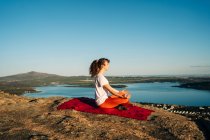 Side view of young lady with long curly hair in activewear meditating with closed eyes in Lotus pose sitting on rocky hill slope above sea under cloudless blue sky — Stock Photo