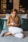Mujer alegre sentada en el sofá y disfrutando de la música en los auriculares mientras mira la pantalla del teléfono inteligente - foto de stock
