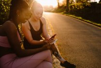 Allegro multirazziale atlete in activewear seduto sulla panchina nel parco e utilizzando i telefoni cellulari insieme dopo l'allenamento al tramonto — Foto stock