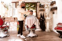 Male hairdresser in textile mask trimming hair of anonymous client in armchair against mirror in barbershop — Photo de stock