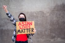 Femme ethnique avec masque et plaque en carton avec inscription Asian Lives Matter protestant avec le bras levé dans la rue de la ville et détournant les yeux — Photo de stock