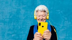 Frohe ältere Frau in gelben Kopfhörern, die Musik hört, während sie auf lebendigem blauen Hintergrund mit dem Handy surft — Stockfoto