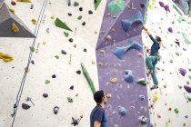 From below of brave female athlete climbing artificial wall in bouldering center under supervision of professional instructor — Stock Photo