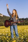 Mujer hipster joven reflexiva de pie en un prado apuntando hacia el cielo en el campo y tocando la guitarra durante la luz del sol de verano - foto de stock