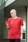 Jovem homossexual fêmea em t-shirt e fones de ouvido com celular olhando para a tela enquanto ouve música — Fotografia de Stock