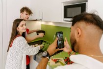 Side view of young ethnic guy using smartphone while taking photo of positive diverse friends cooking together in kitchen — Foto stock