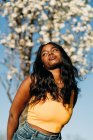 Низкий угол красивой афроамериканской женщины, стоящей в цветущем весеннем парке и наслаждающейся солнечной погодой, смотрящей в сторону — стоковое фото