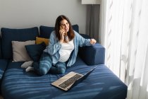 Pregnant female freelancer sitting on sofa with laptop in living room and speaking on mobile phone while discussing remote project and working from home — Stock Photo
