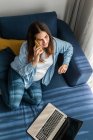 Freelancer embarazada sentada en un sofá con portátil en la sala de estar y hablando por teléfono móvil mientras trabaja desde casa - foto de stock