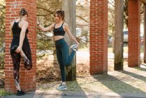 Sportswoman with anonymous girlfriend in activewear exercising with raised legs on pavement in town on sunny day — Stock Photo