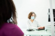 Обрезанная неузнаваемая афроамериканка во время приема сидит у врача в маске для лица за столом, защищенным стеклянным экраном в современной клинике во время вспышки коронавируса — стоковое фото