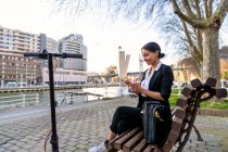 Lächelnde ethnische Unternehmerin sitzt mit überkreuzten Beinen auf einer Holzbank und telefoniert, während sie auf einer Bank in der Stadt gegen einen Roller wegschaut — Stockfoto