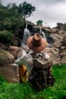 Назад перегляд анонімний чоловічий рюкзак у капелюсі, насолоджуючись видом каскадного потоку з грубого каменю у виправдувальній природі — стокове фото